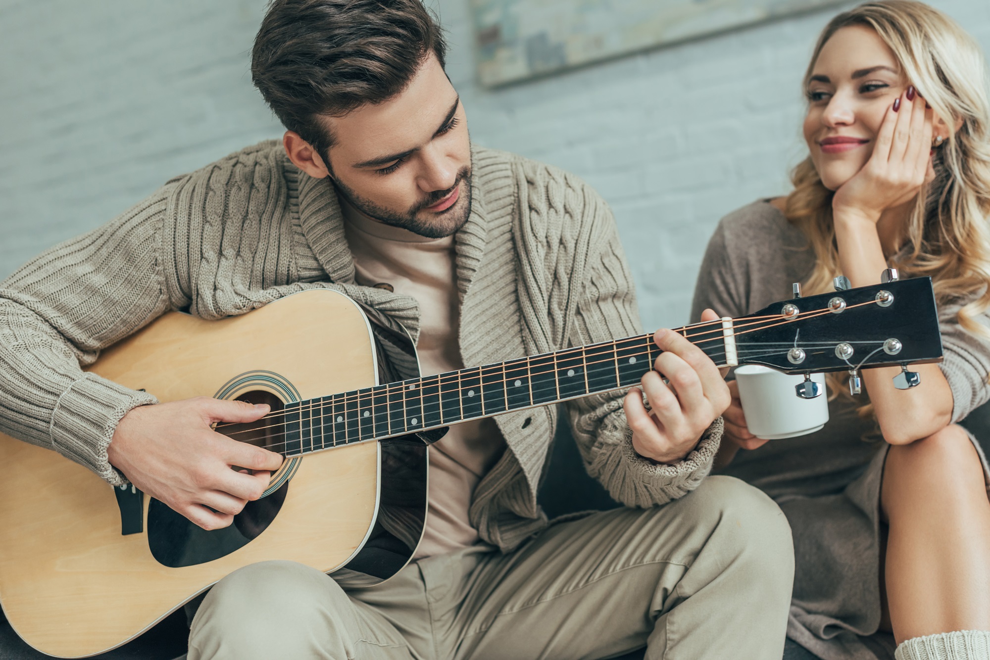 handsome young man playing guitar for girlfriend at home while she sitting on couch with coffee